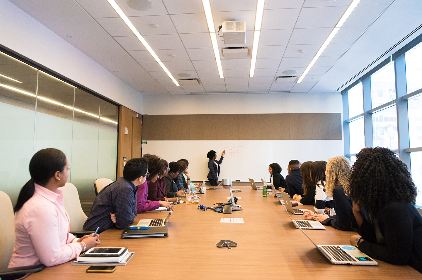 photo of people gathered around a meeting room table with a person presenting at a white board at the far end of the room