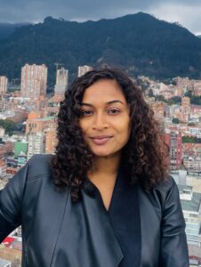 Individual smiling at the camera with a backdrop of a densely built city and green mountains, under a cloudy sky.