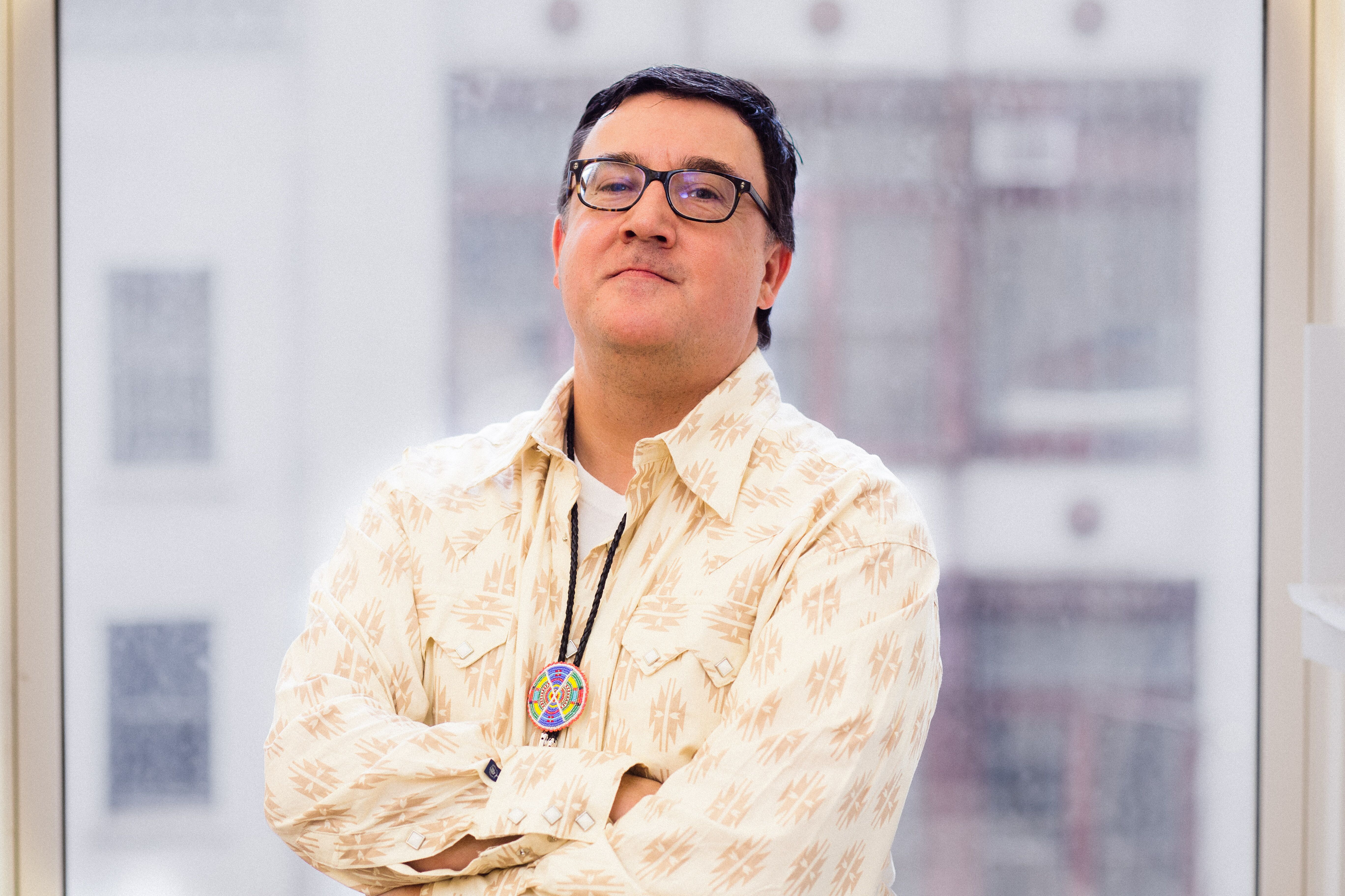 Person in a patterned shirt and glasses, wearing a lanyard with a colorful pendant, stands in front of a window with a cityscape in the background.