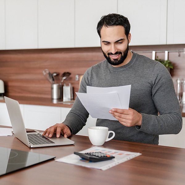 Man in his kitchen, looking at a paper, with a calculator and laptop in view